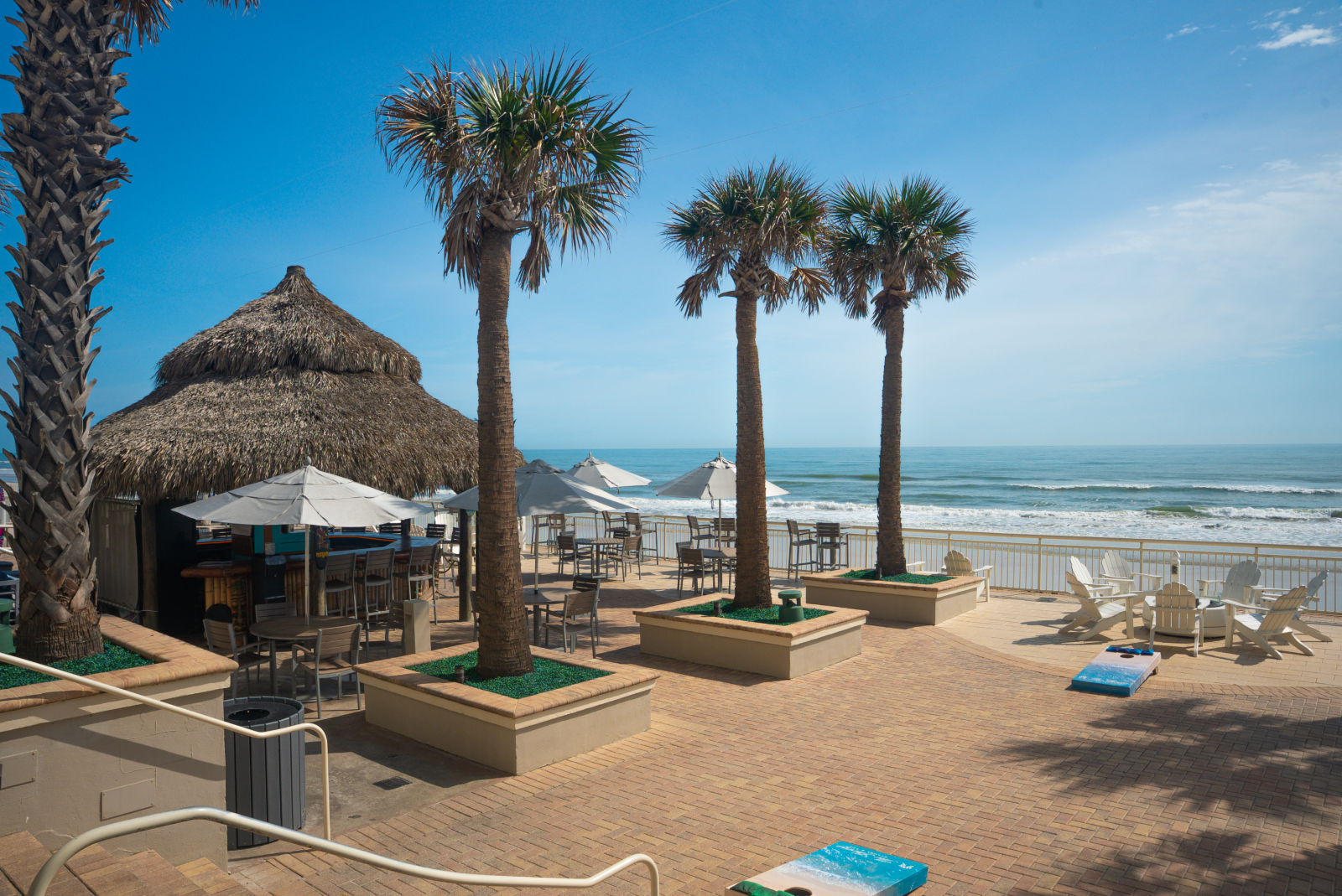 A peaceful beach scene with palm trees, umbrellas, chairs, a thatched hut, and ocean waves under a blue sky.