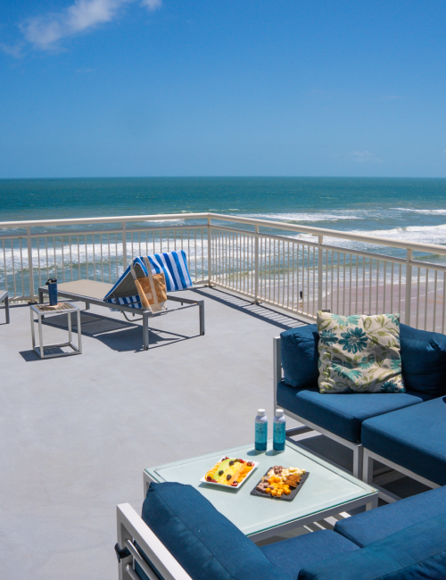 A balcony with blue seating, towels, drinks, snacks, overlooking the ocean with waves and a partly cloudy sky.