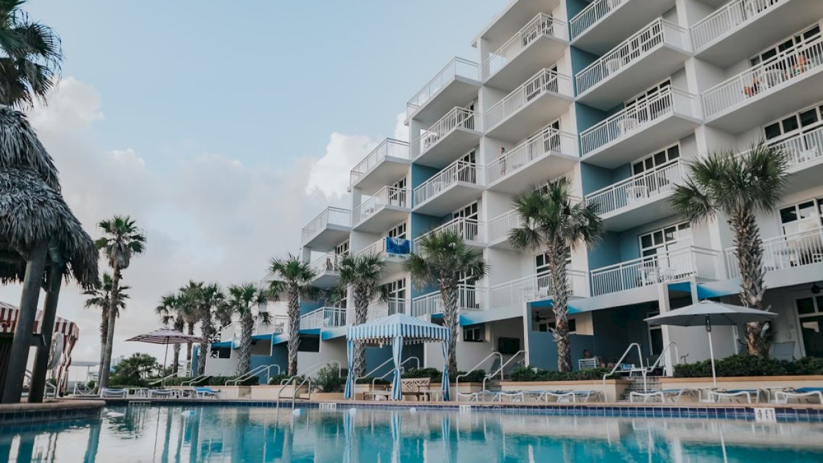 The image shows a multi-story resort building with balconies, surrounded by palm trees and a pool in the foreground.