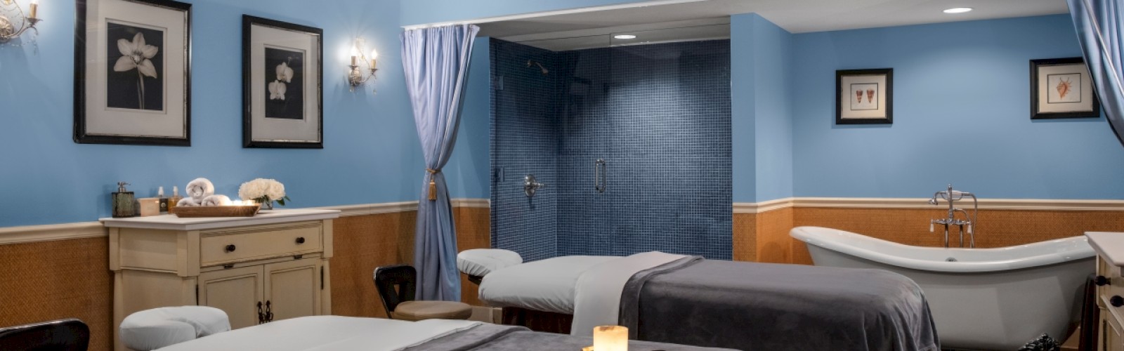 This image shows a spa treatment room with two massage tables, a bathtub, candles, and wall art, creating a relaxing ambiance.