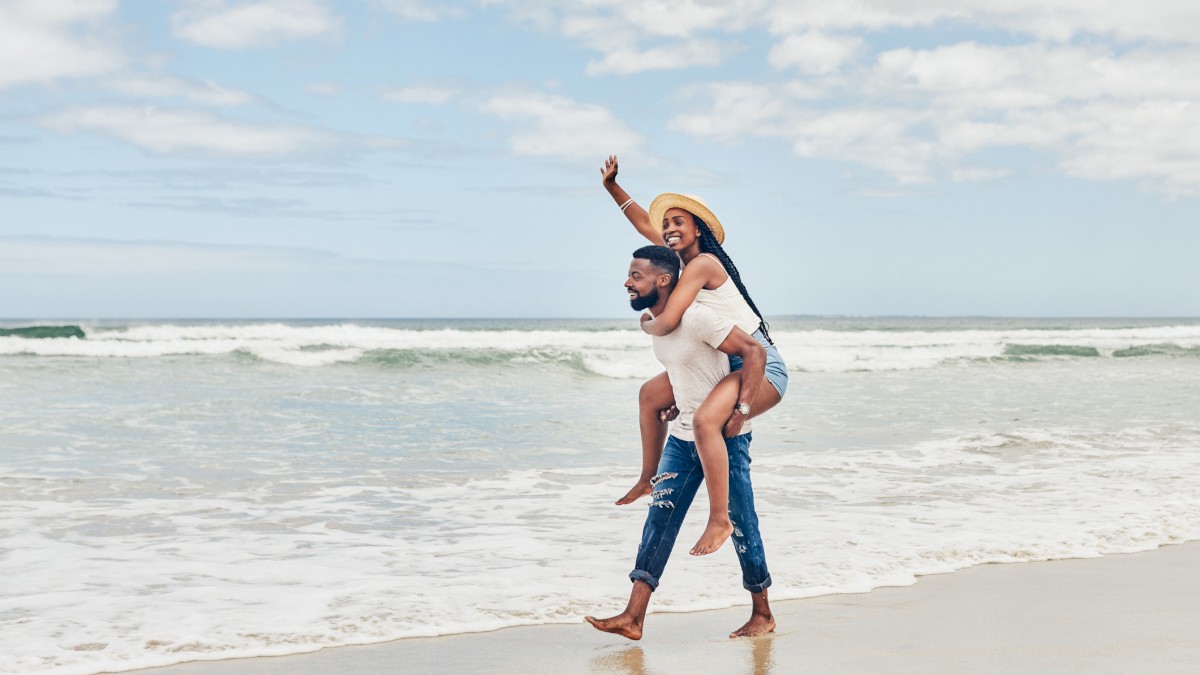A couple enjoys a playful piggyback ride on a sandy beach with ocean waves in the background, under a partly cloudy sky.