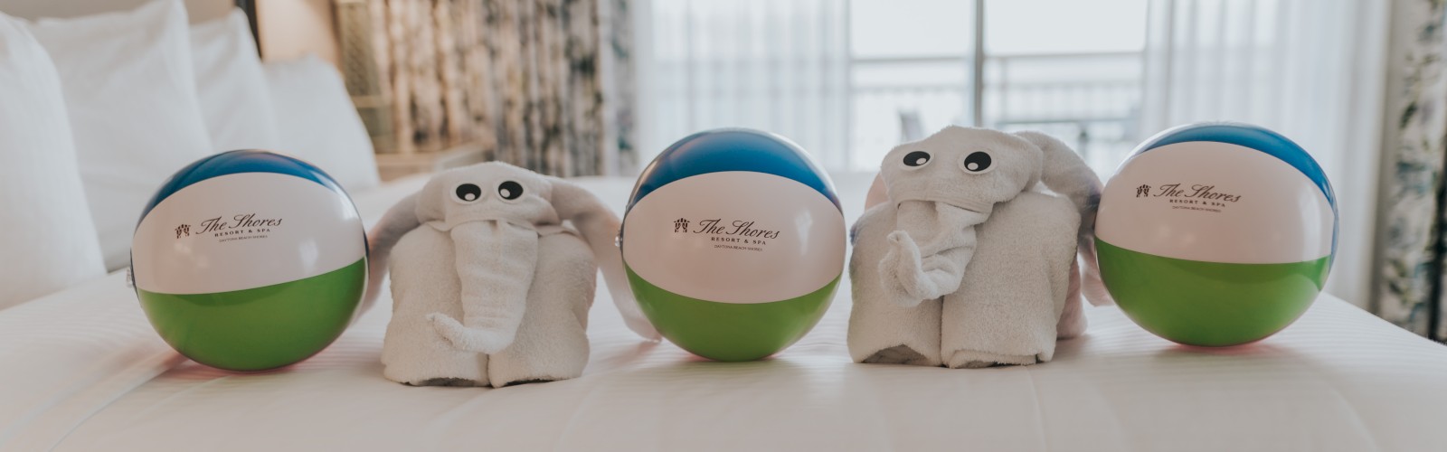 Beach balls with towel elephants are arranged on a neatly made bed by a window in a cozy room.