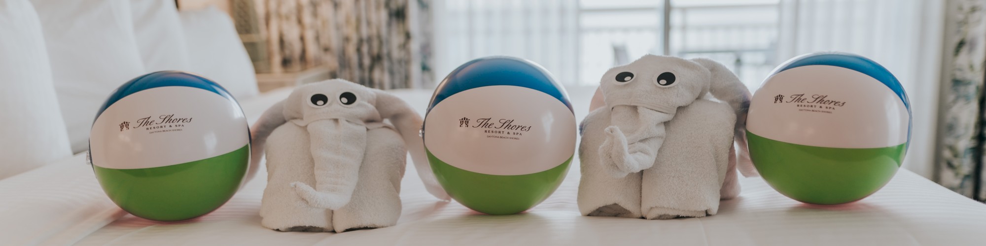 Beach balls with towel elephants are arranged on a neatly made bed by a window in a cozy room.