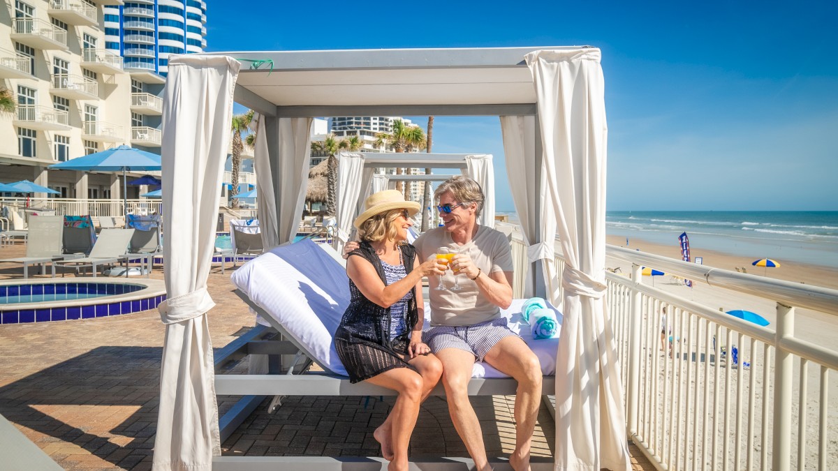 Two people sit under a beach cabana, toasting drinks, with buildings, a pool, and the ocean in the background on a sunny day.