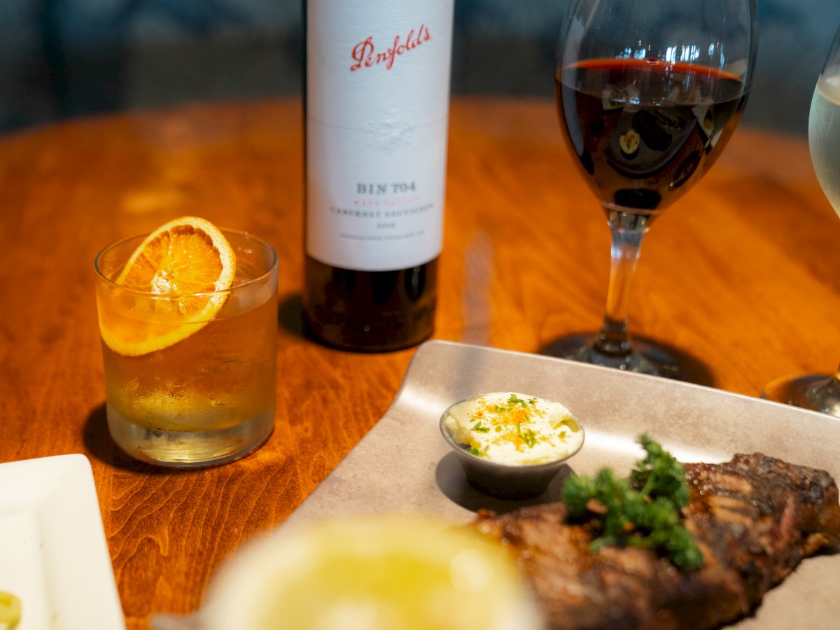 A table with a steak, glass of red wine, cocktail with an orange slice, Penfolds wine bottle, and a side of sauce.