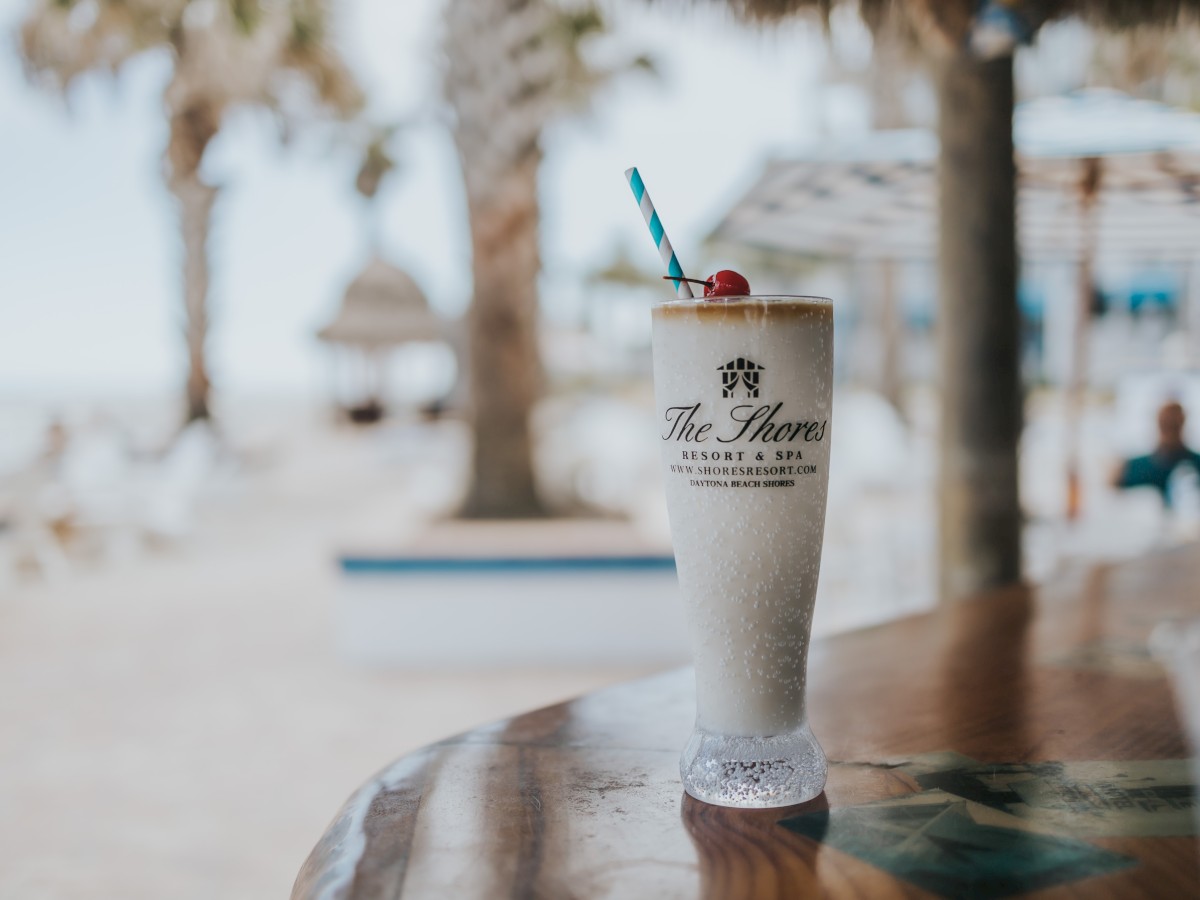 A milkshake in a tall glass with a cherry on top on a wooden table by the beach, palm trees in the background.