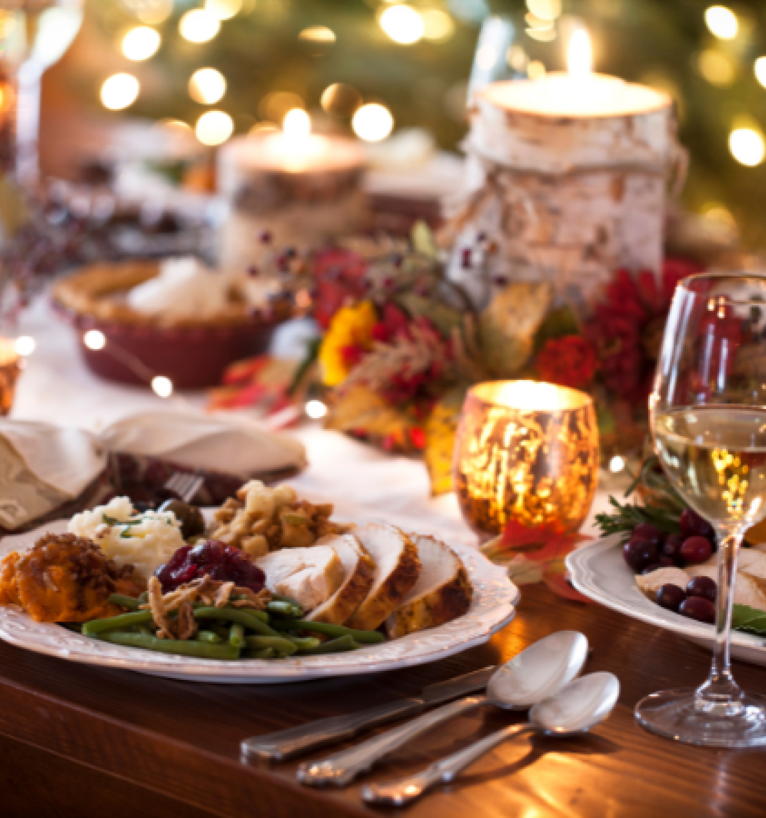 A festive dinner table with candles, wine glasses, and plates of food, including sliced turkey and vegetables in a warm, cozy setting.