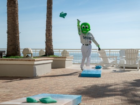 A person in a green mascot costume is playing cornhole outdoors near palm trees and a body of water.