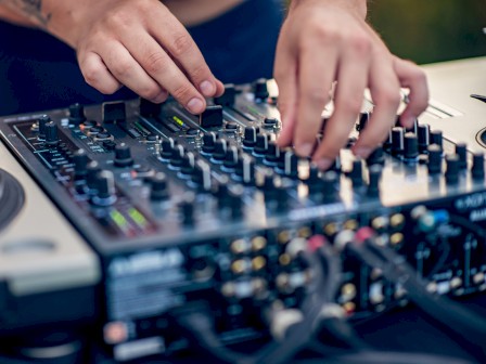 A DJ is adjusting controls on a sound mixer, surrounded by equipment like turntables, suggesting a music mixing setup outdoors.