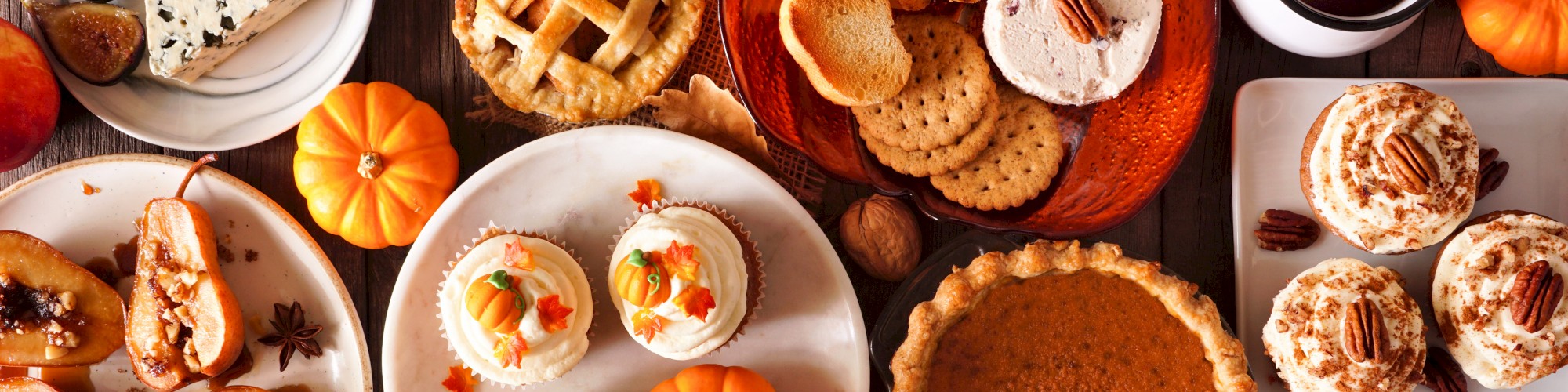 Assorted autumn foods: pies, muffins, cheese, apples, figs, pumpkin, biscuits, and spiced drink on a wood table.