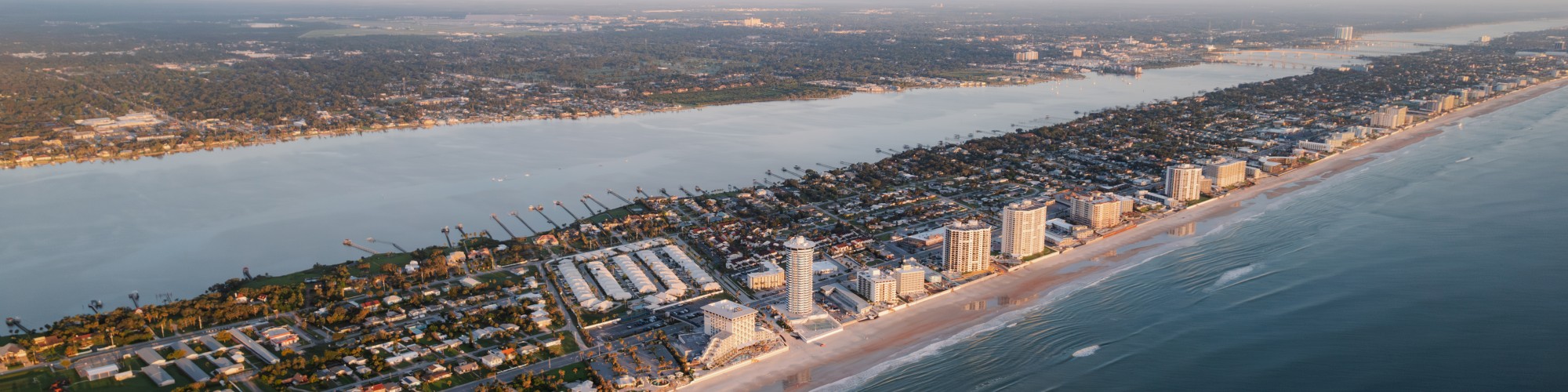 Aerial view of a coastal city with sandy beaches, tall buildings, and a body of water on both sides, captured at dawn or dusk.