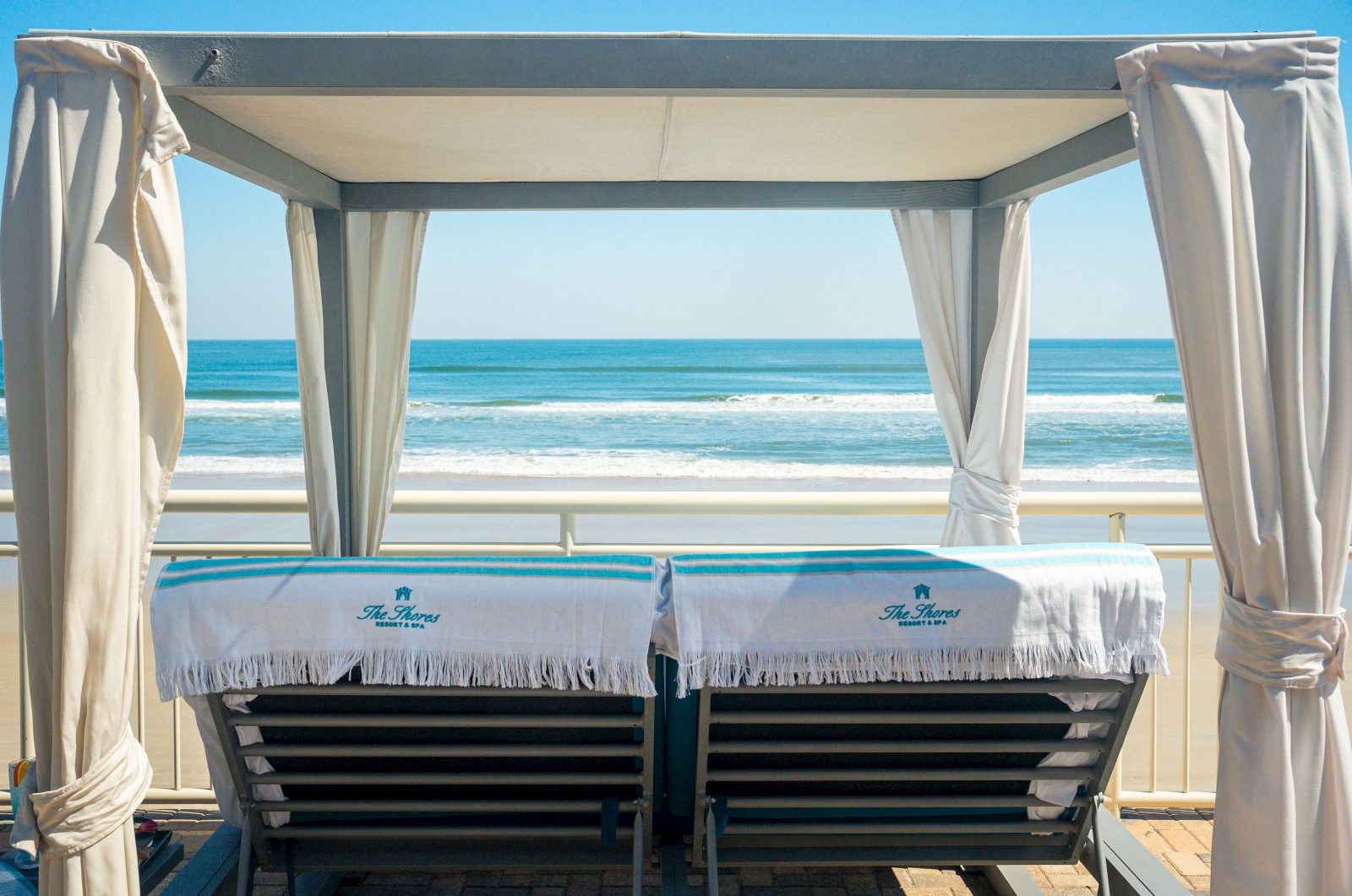 Two lounge chairs under a canopy face the ocean, with towels draped over them, offering a serene beach view under blue skies.