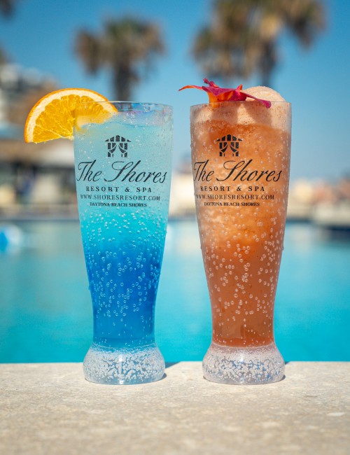 Two colorful drinks served in tall glasses by a poolside, each garnished with fruit, branded with 