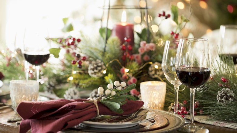 A festive table setting with wine glasses, candles, a decorative greenery centerpiece, and red napkins.