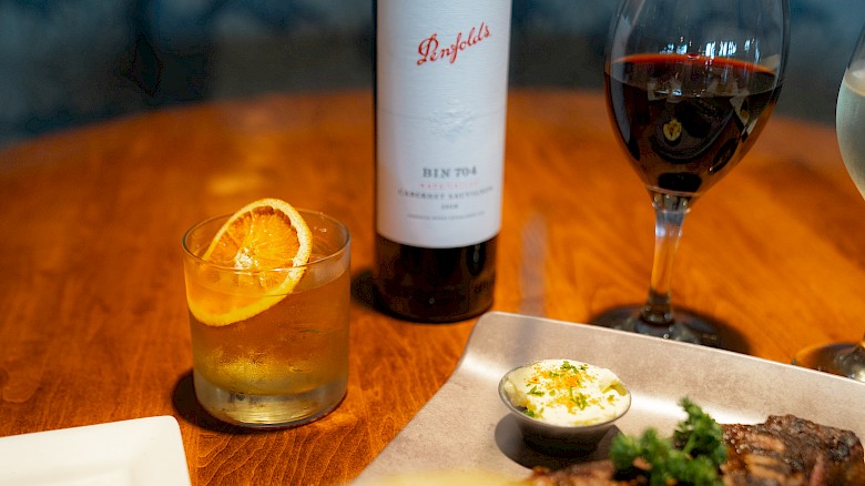 A table set with wine, a cocktail garnished with an orange slice, steak with sauce, and water on a wooden surface.