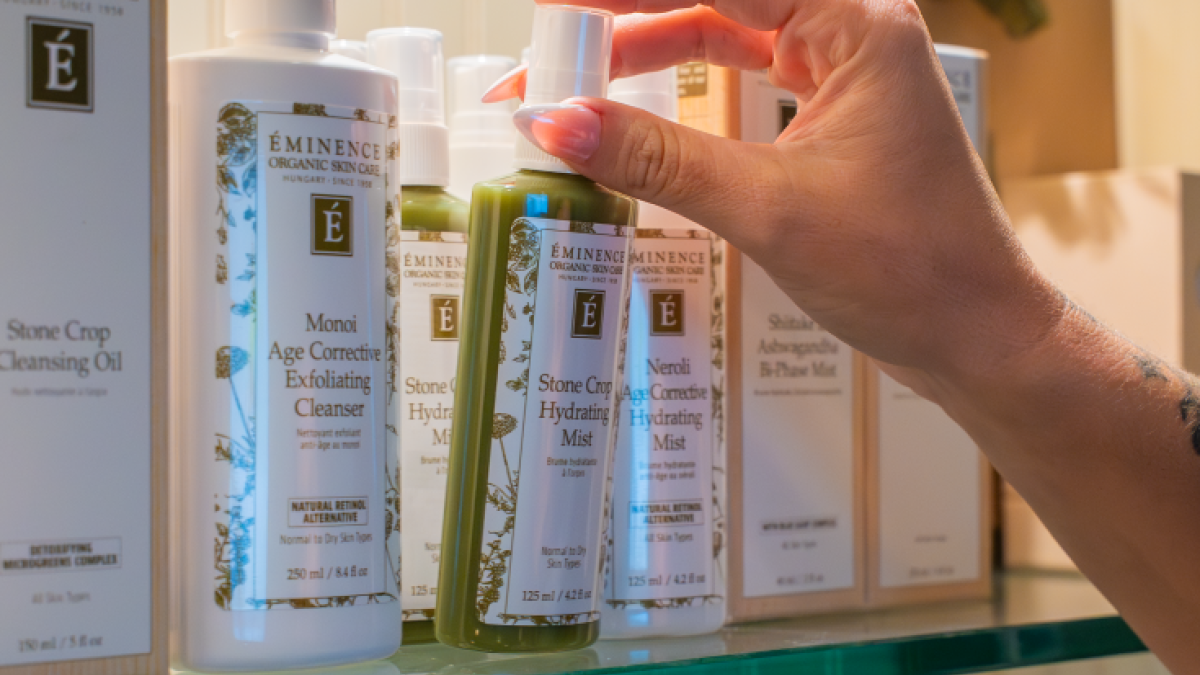 The image features a hand spritzing a green bottle of skincare mist from Eminence Organics on a glass shelf, with other skincare products nearby.