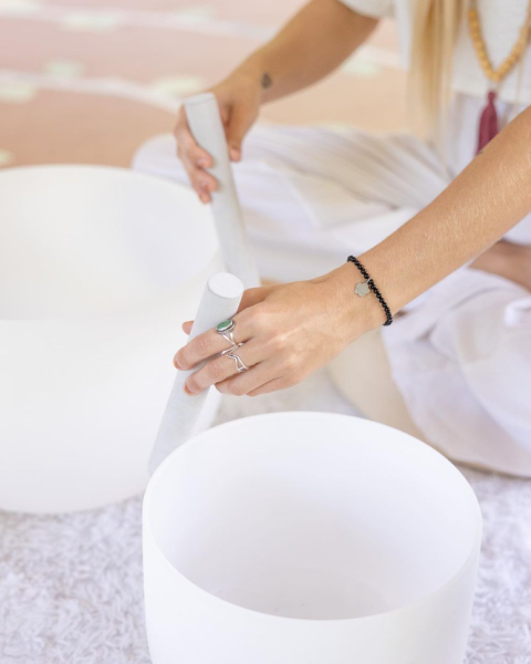 A person is playing crystal singing bowls with a mallet while seated on the floor, dressed in white clothing, and wearing jewelry.