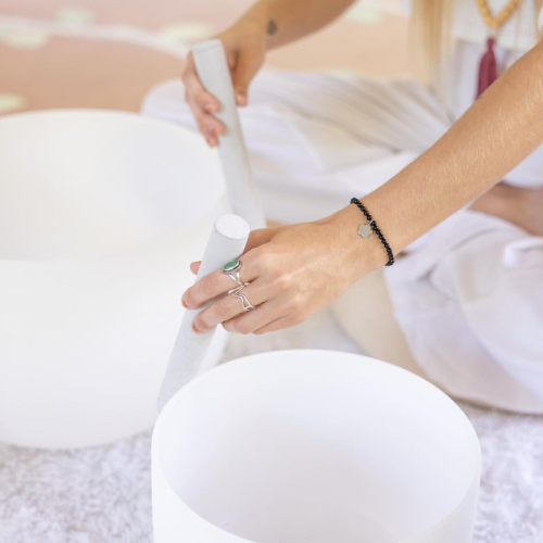 A person is playing crystal singing bowls with a mallet while seated on the floor, dressed in white clothing, and wearing jewelry.