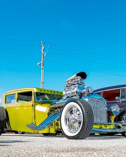 The image shows two vintage hot rods parked under a clear blue sky, one green and one dark red, with visible chrome detailing and large engines.