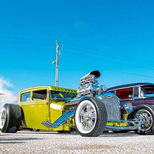 The image shows two vintage hot rods parked under a clear blue sky, one green and one dark red, with visible chrome detailing and large engines.