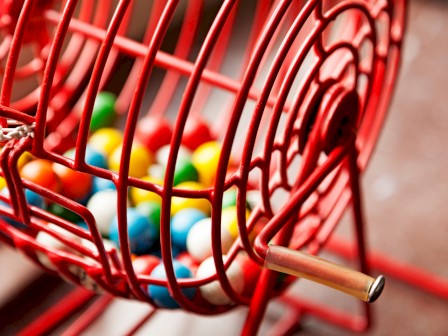 The image shows a red bingo cage filled with colorful balls, featuring a crank handle for rotating the cage to select balls.