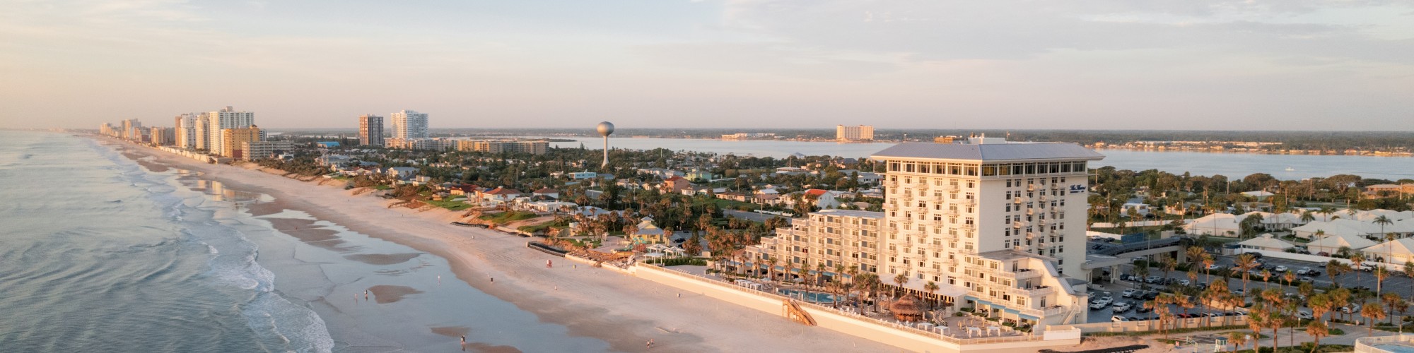 Aerial view of a beach with gentle waves, buildings, and a coastal city in the distance, under a clear sky.