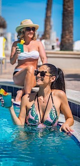 Two women enjoying drinks by the pool, one in the water and the other sitting on the edge, with palm trees in the background.