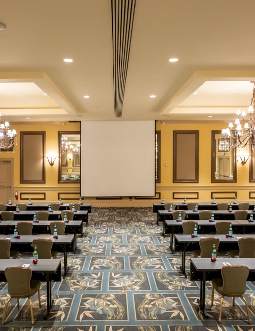 A conference room with rows of tables and chairs, a large screen, chandeliers, and decorative mirrors on the walls.