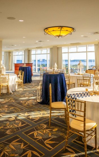 A banquet room with round tables draped in blue and white linens, golden chairs, and large windows offering a scenic view outside.