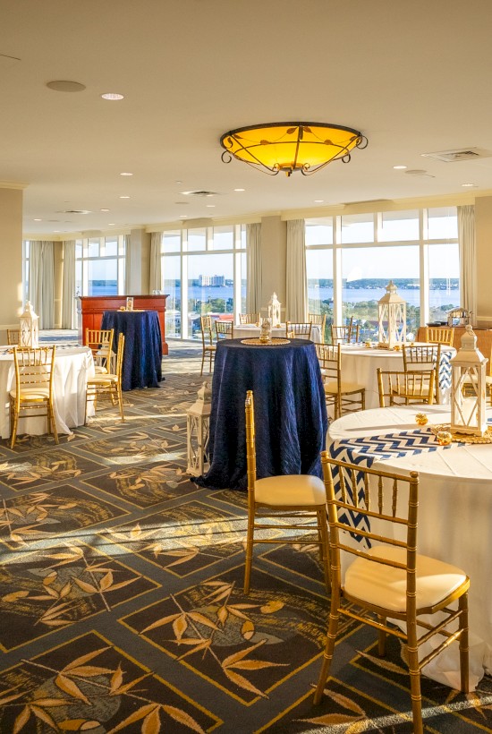 A banquet room with round tables draped in blue and white linens, golden chairs, and large windows offering a scenic view outside.