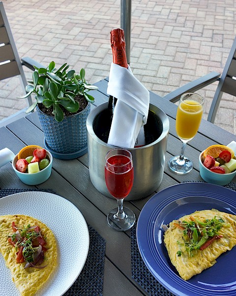 An outdoor table with two omelettes, fruit bowls, champagne, and juice, set with napkins and a plant centerpiece.