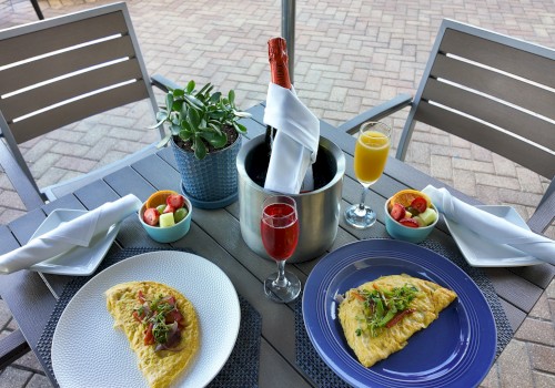 An outdoor table with two omelettes, fruit bowls, champagne, and juice, set with napkins and a plant centerpiece.