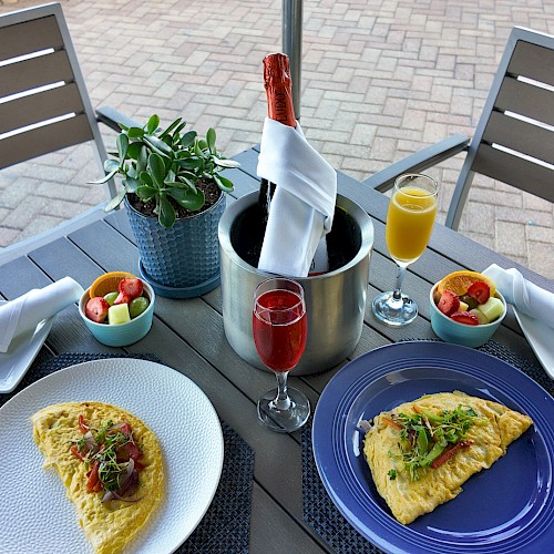 An outdoor table with two omelettes, fruit bowls, champagne, and juice, set with napkins and a plant centerpiece.