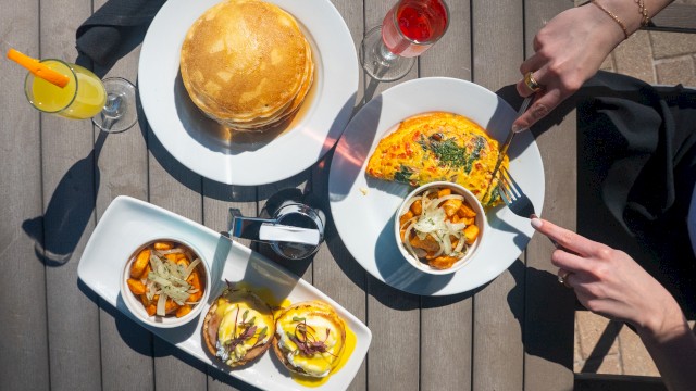 A brunch setting with pancakes, an omelette, Benedicts, drinks, and hash browns on a wooden table, with hands holding utensils.
