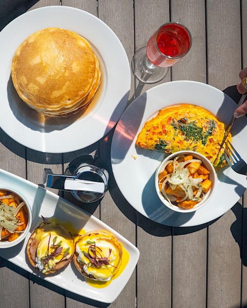 A top-down view of a brunch: two plates with pancakes and an omelette, a small dish of veggies, and burgers with fries, plus drinks on a wooden table.