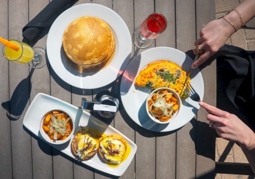 A top-down view of a brunch: two plates with pancakes and an omelette, a small dish of veggies, and burgers with fries, plus drinks on a wooden table.