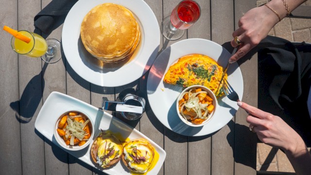 A top-down view of a brunch: two plates with pancakes and an omelette, a small dish of veggies, and burgers with fries, plus drinks on a wooden table.