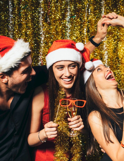 A group of people celebrating, wearing Santa hats and holding drinks, with a festive gold backdrop. Everyone looks happy and engaged in fun.