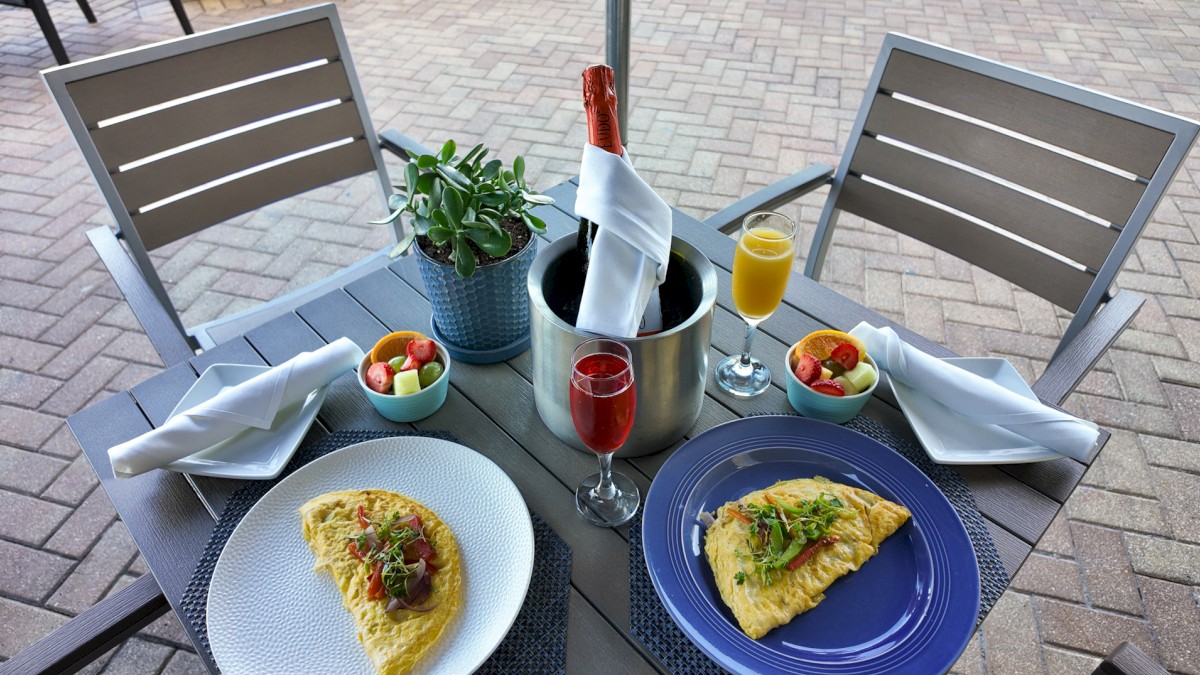 The image shows a patio table with two omelets, fruit bowls, drinks, and a champagne bottle in a bucket, set for an outdoor meal.