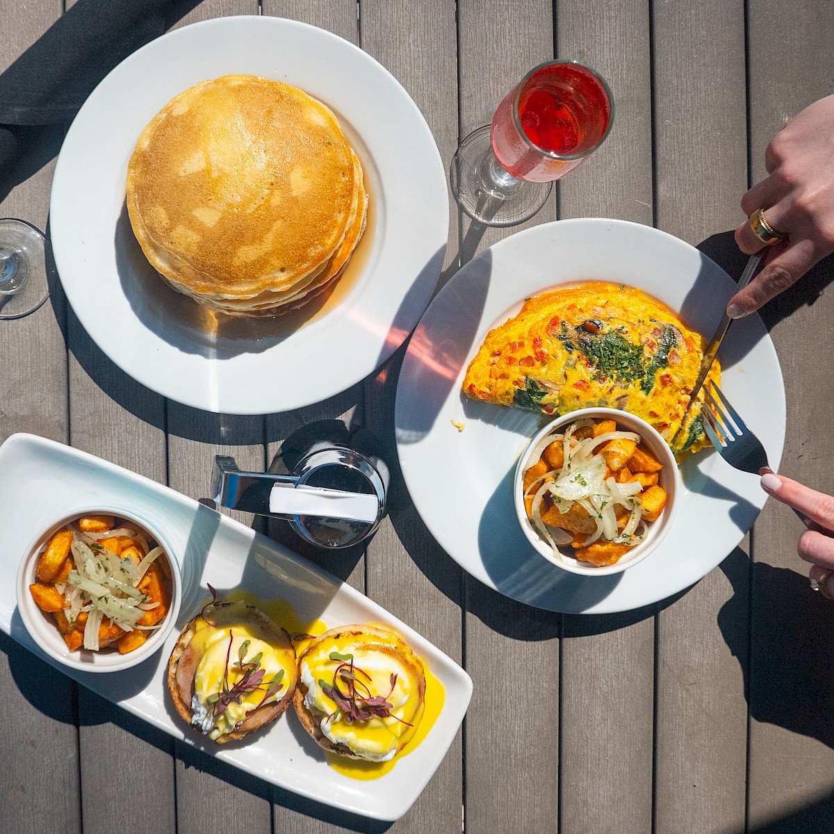A breakfast spread featuring pancakes, an omelet, Eggs Benedict, home fries, a mimosa, and a red drink on a wooden table.