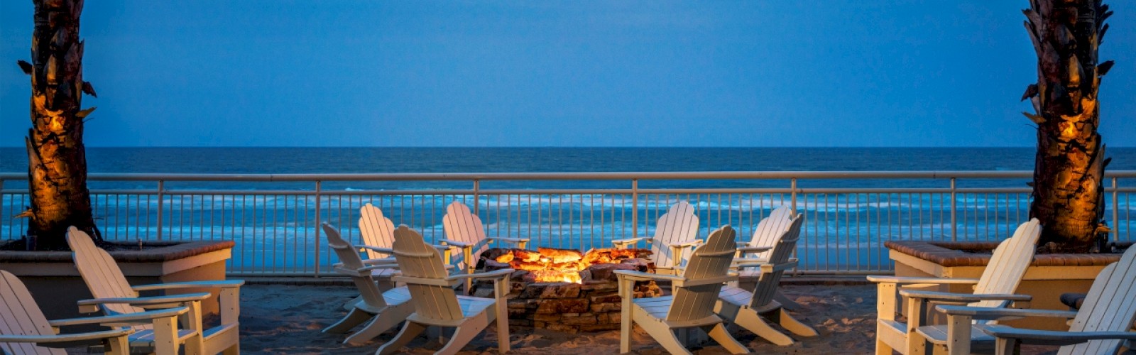 A cozy outdoor seating area with a fire pit, surrounded by Adirondack chairs, overlooking the ocean under a twilight sky, framed by palm trees.
