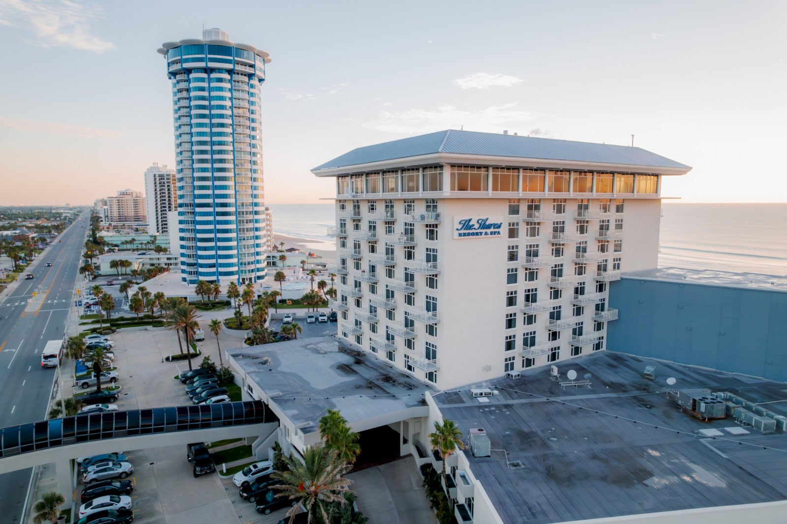 A coastal cityscape with a tall modern building, a hotel, palm trees, and cars. The ocean is visible in the background, under a clear sky.