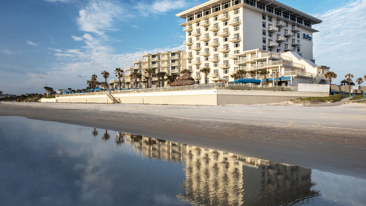 A beachfront hotel with several balconies reflects in the water, surrounded by palm trees under a partly cloudy sky.