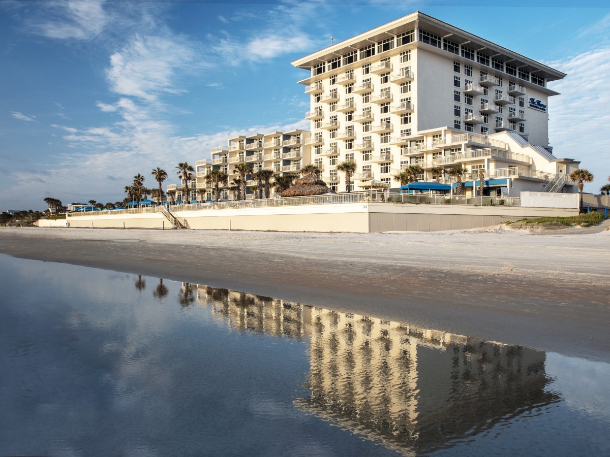 A beachfront hotel with several balconies reflects in the water, surrounded by palm trees under a partly cloudy sky.