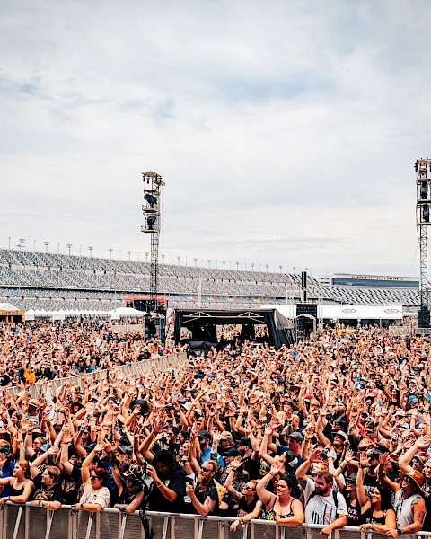 A large outdoor concert crowd is gathered in front of a stage, with stadium seating visible in the background under a partly cloudy sky.