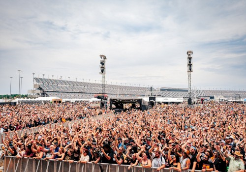 A large outdoor concert crowd is gathered in front of a stage, with stadium seating visible in the background under a partly cloudy sky.