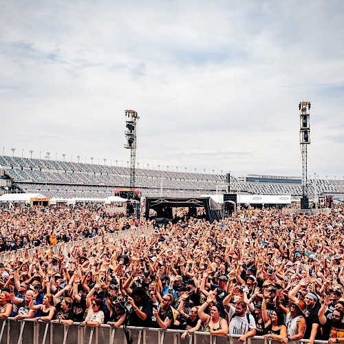 A large outdoor concert crowd is gathered in front of a stage, with stadium seating visible in the background under a partly cloudy sky.