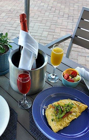 A table with two omelettes, fruit bowls, champagne, and orange juice set for a meal outdoors, with napkins and a plant.