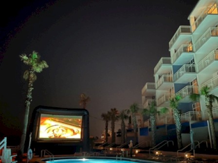 A nighttime poolside movie screening with a large inflatable screen and an adjacent multi-story building.