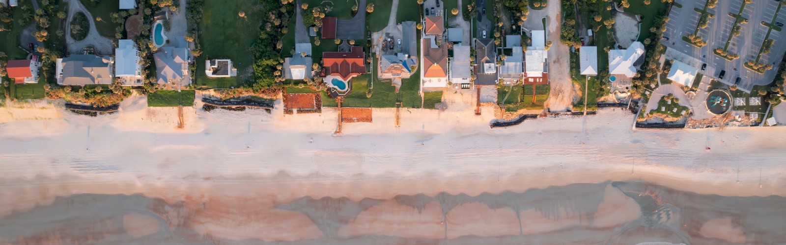 Aerial view of a beachfront neighborhood with houses, a road, sandy beach, and ocean waves hitting the shore.